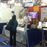 Two employees talking to a business woman while standing behind a trade show booth for the Engineering Innovation product Confirm Delivery.