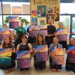 Group of Engineering Innovation employees in a painting studio, each holding a painting of the ocean during sunset.