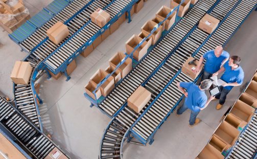 Aerial view of a factory floor with three workers in blue shirts discussing beside conveyor belts carrying cardboard boxes. Despite the efficient packaging process, the scene subtly hints at a labor shortage with only a few employees managing the tasks along the parallel lines.
