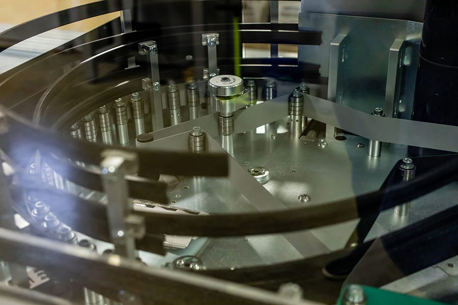 Close-up of industrial machinery with metallic components, including a circular assembly and multiple rods and belts. The setup suggests a mechanical process or assembly line in a factory setting.