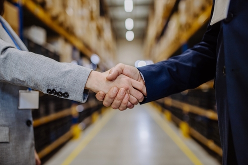 Two people shaking hands in a warehouse aisle, surrounded by shelves filled with boxes. Both are dressed in business attire, with one wearing a light gray suit and the other a dark blue suit. The background is slightly blurred.