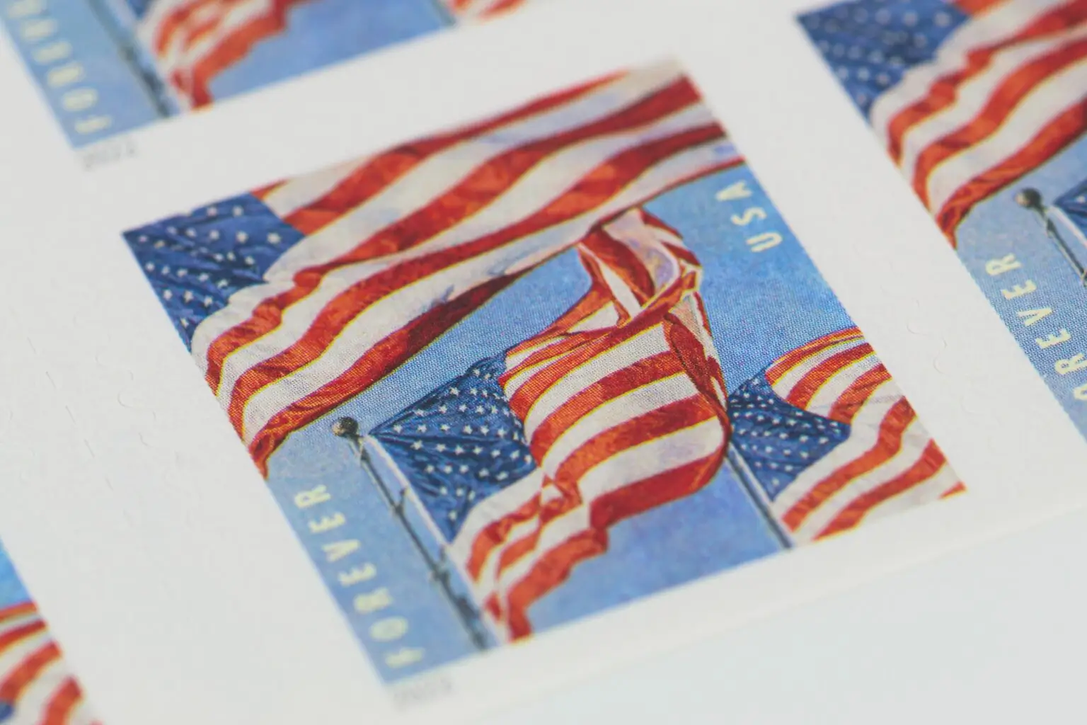 Close-up of U.S. postage stamps featuring the American flag waving in the wind against a blue sky. The word "FOREVER" and "USA" are visible alongside the flags.