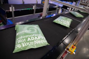 Green packages labeled "Automation That ADAPTS To Your Needs" move along a black conveyor belt in an industrial setting. The system appears to be automated and efficient.