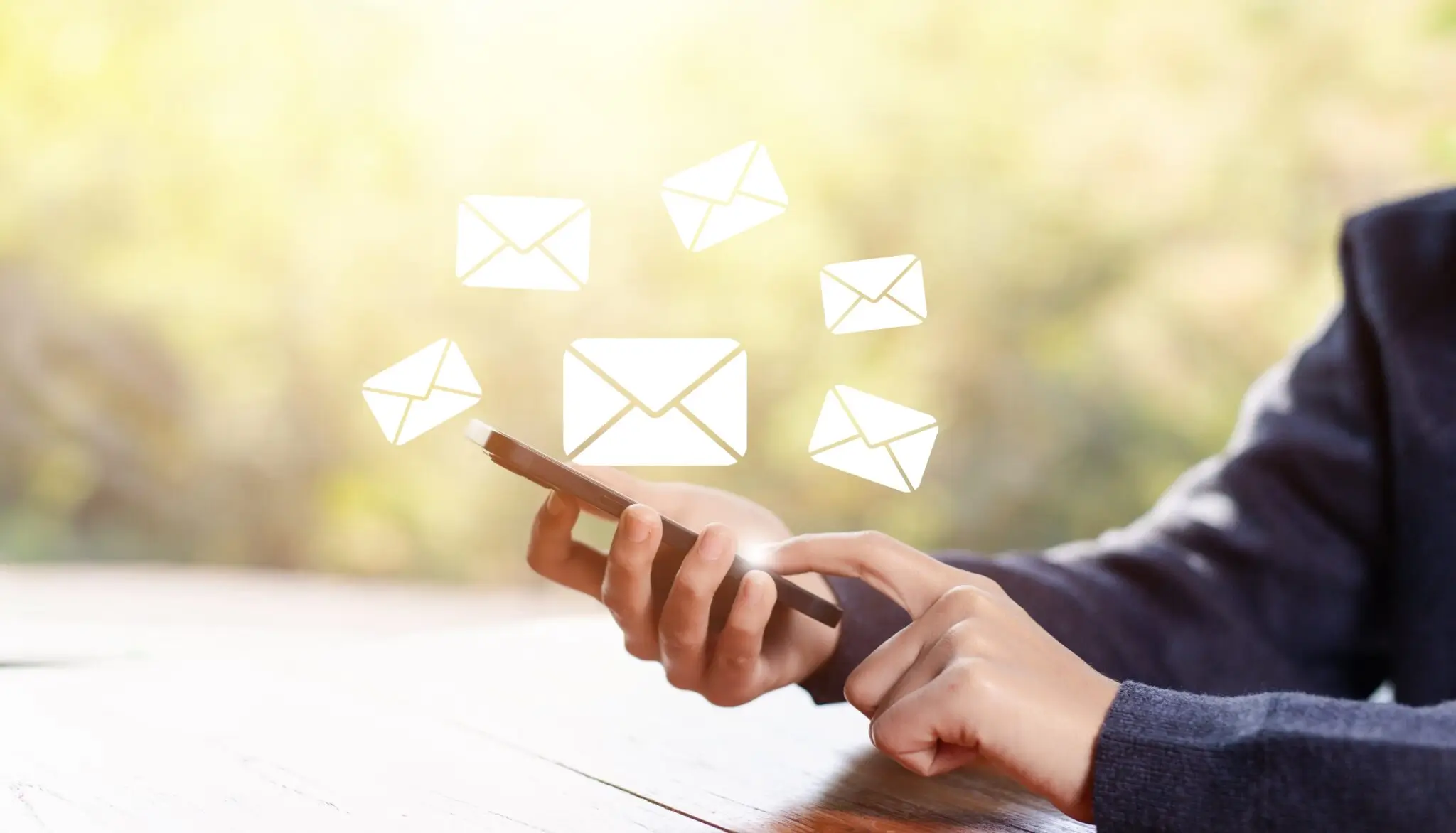 A person is using a smartphone at a wooden table, with digital icons of envelopes floating above, symbolizing sending or receiving emails. The background is softly blurred with natural lighting.