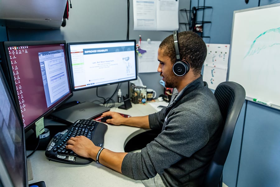 A person wearing headphones sits at a desk with three monitors, focusing on work. The screens display documents and a graph. The workspace is organized with a keyboard, mouse, and a whiteboard in the background.