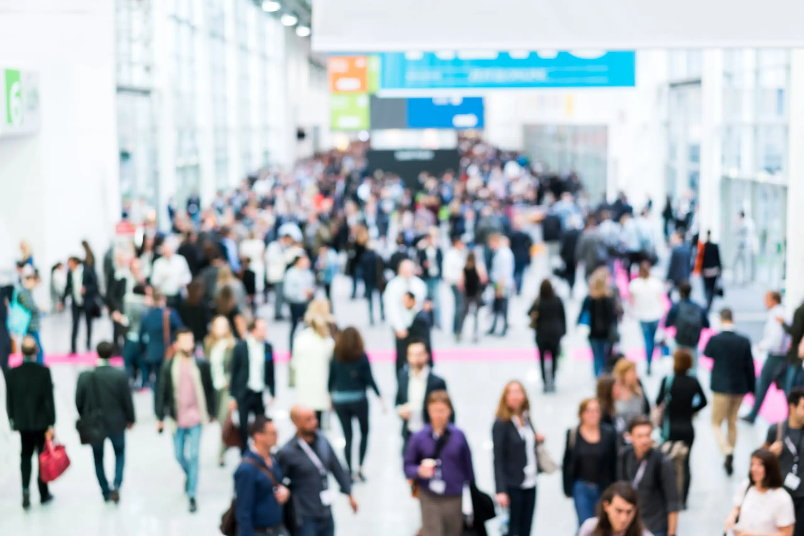 A crowded convention center hallway filled with many people walking in various directions. The scene is bustling, with individuals wearing a mix of business and casual attire, and banners hanging in the background. The focus is blurry.