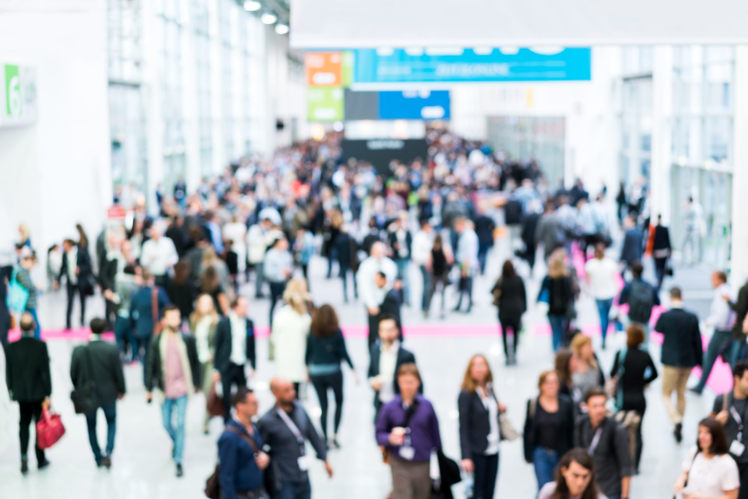 A large crowd of people walking through a busy convention center with bright lighting and signage overhead. The setting has a lively atmosphere, and the crowd consists of diverse individuals engaged in conversation and movement.