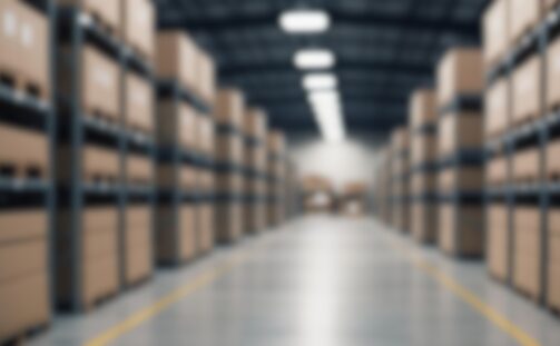 Blurred image of a warehouse with rows of shelves stacked with cardboard boxes. The floor is smooth, and lights illuminate the space from above, creating a vanishing point perspective down the aisle.