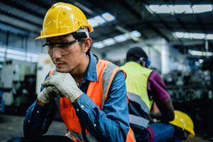 A male factory worker wearing a yellow hard hat, safety glasses, gloves, and an orange vest sits with a thoughtful expression, while another worker in a safety vest sits in the background in an industrial setting.