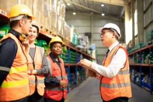 Four workers in safety vests and hard hats stand in a warehouse aisle. One person holds a clipboard and appears to be instructing the others, who listen attentively. Shelves stocked with boxes line the warehouse.