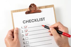 A person holds a clipboard with a checklist. They are checking off items using a red pen, with the first three boxes checked and the remaining boxes empty.