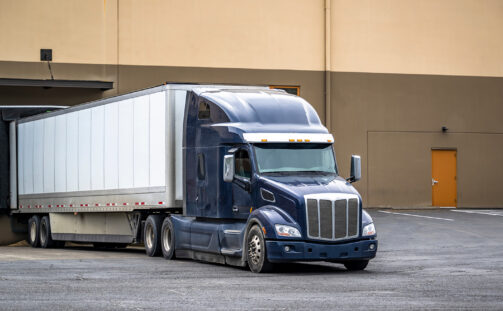 A large blue semi-truck with a white trailer is backed up to a loading dock at a warehouse building, preparing to load or unload cargo. The area around the building appears empty.