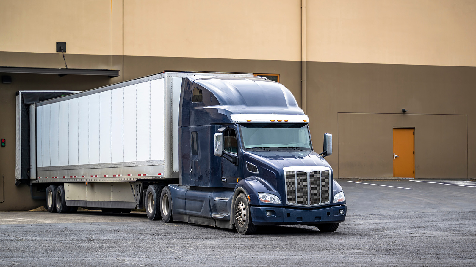 A large blue semi-truck with a white trailer is backed up to a loading dock at a warehouse building, preparing to load or unload cargo. The area around the building appears empty.