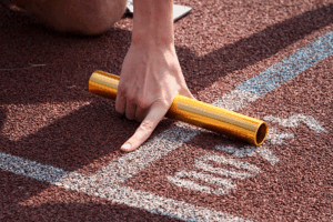 Close-up of a person's hand holding a gold relay baton, positioned on the starting line of a running track. The hand points to the track, indicating readiness for a relay race.