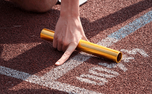 Close-up of a person's hand holding a gold relay baton, positioned on the starting line of a running track. The hand points to the track, indicating readiness for a relay race.