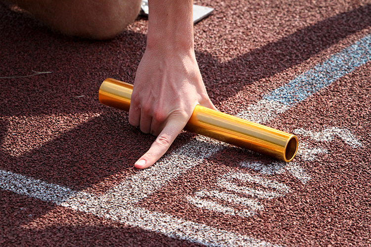 Close-up of a person's hand holding a gold relay baton, positioned on the starting line of a running track. The hand points to the track, indicating readiness for a relay race.