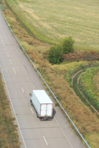 A white box truck drives alone on a rural, empty road bordered by grassy fields and greenery on a clear day, viewed from above.