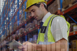 A man in a yellow safety vest and helmet checks a clipboard in a warehouse aisle, with shelves full of boxes and overlaid graphics of data charts and graphs.