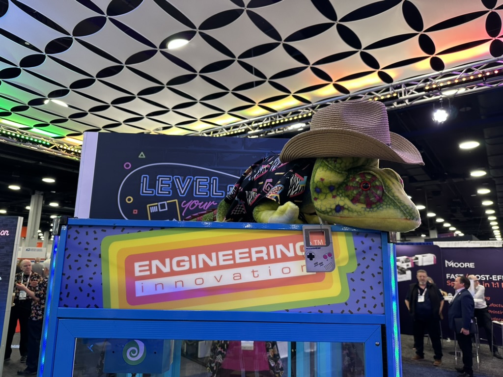 A large chameleon statue wearing a cowboy hat and jacket sits atop a colorful "Engineering Innovation" booth at a convention. The ceiling has a geometric pattern and people are visible in the background.