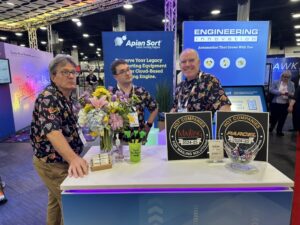 Three men in matching patterned shirts stand behind a booth at a trade show. The booth displays flowers, awards, and promotional signs for engineering and cloud-based mailing solutions. Bright lights and screens are in the background.