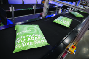 Green packages labeled “Automation That ADAPTS To Your Needs” move along a black conveyor belt in an industrial setting, likely representing automated packaging or sorting technology.
