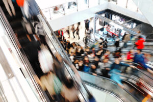 A busy shopping mall interior with crowds of people riding escalators and walking around, creating a sense of movement and bustling activity. The scene is brightly lit with a modern design.
