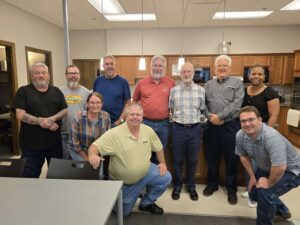 A group of ten adults, men and women, stand and kneel together in a kitchen or break room, smiling at the camera. The room has wooden cabinets, pendant lights, and tables and chairs in the foreground.