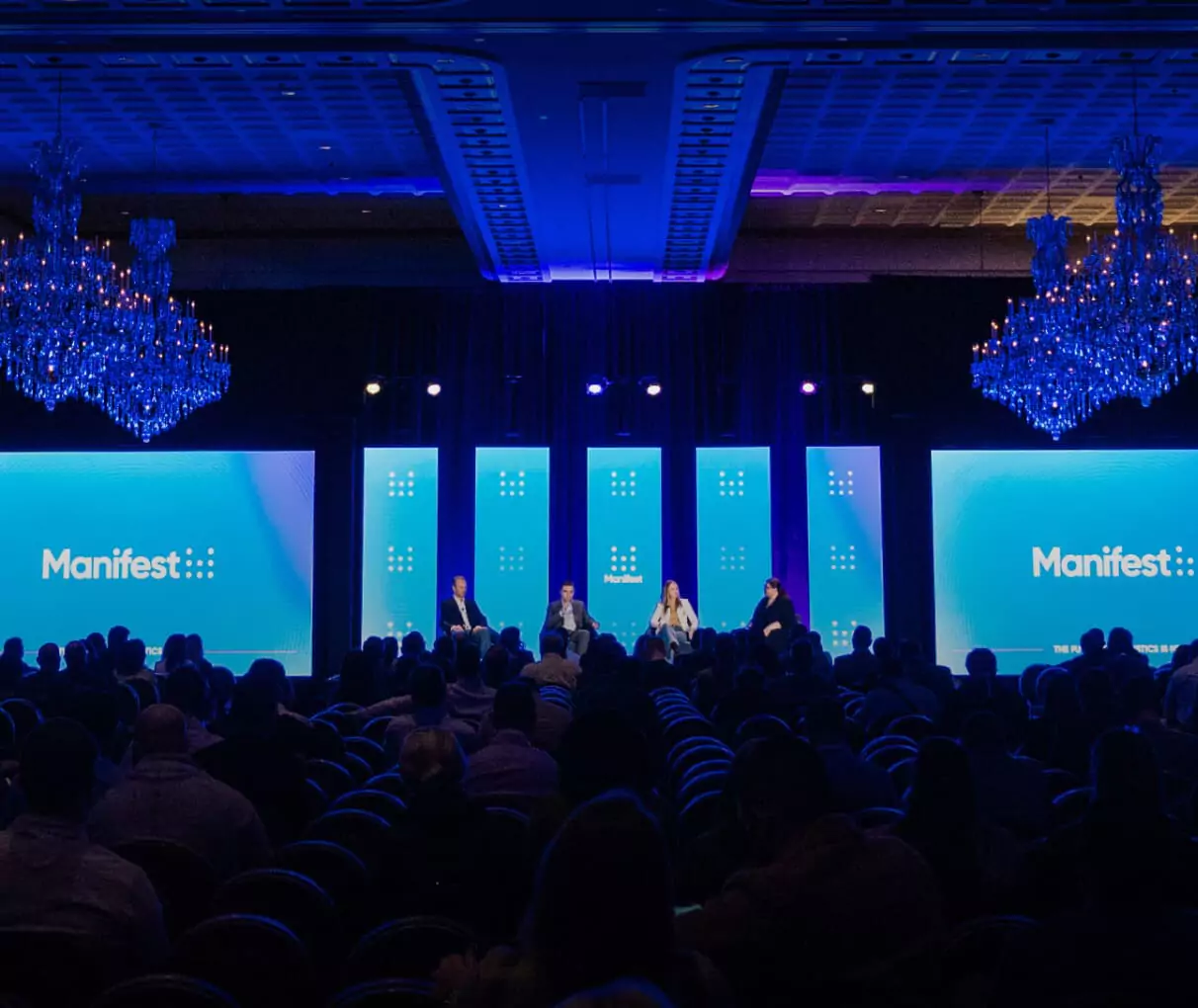 A panel of five people sits onstage at a conference called "Manifest." The audience faces the stage, which is lit by blue and purple lights, with large screens displaying the event name on both sides.