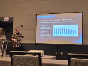 A person stands near a podium, presenting a slide titled "The New Warehouse Landscape" to an audience. The slide discusses robotics and automation adoption trends with a chart displayed on a projector screen.