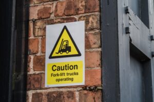 A caution sign on a brick wall shows a black forklift icon inside a yellow triangle above the text: "Caution Fork-lift trucks operating.