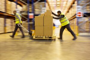 Two warehouse workers in yellow safety vests move boxes on pallets through a large warehouse aisle lined with tall shelves filled with goods. The image shows motion blur, emphasizing the workers’ movement.