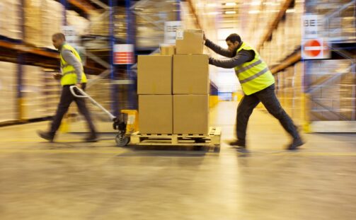 Two warehouse workers in yellow safety vests move boxes on pallets through a large warehouse aisle lined with tall shelves filled with goods. The image shows motion blur, emphasizing the workers’ movement.