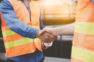 Two people wearing orange safety vests shake hands outdoors, suggesting a construction or work site agreement or partnership. Only their torsos and arms are visible.
