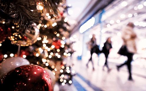 A decorated Christmas tree with red and gold ornaments and twinkling lights in focus, while people walk and shop in a brightly lit indoor mall blurred in the background.