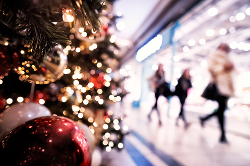 A decorated Christmas tree with red and gold ornaments and twinkling lights in focus, while people walk and shop in a brightly lit indoor mall blurred in the background.