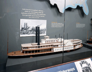 A detailed model of a historic paddlewheel steamboat is displayed in a glass case at a museum, with an old photograph and informational text on the wall behind it.