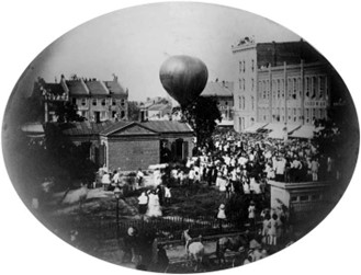 A vintage black-and-white photo shows a large crowd gathered outdoors around a hot air balloon preparing for liftoff in a city square, with historic buildings in the background.