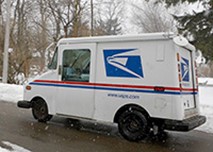 A USPS mail delivery truck drives down a snowy residential street lined with bare trees and snow-covered ground.