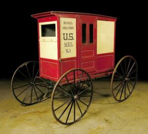 A vintage red and white horse-drawn mail wagon with large spoked wheels and a sign reading "Rural Delivery U.S. Mail No. 1" on the side, displayed indoors against a black background.