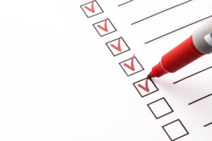 A red marker checks off boxes on a list, filling in red checkmarks on a row of empty and completed checkboxes on white paper.