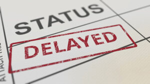 A close-up of a document with the word "STATUS" and a bold red stamp that says "DELAYED" across it, indicating a postponed or rescheduled status.