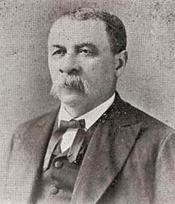 Black-and-white portrait of an older man with short hair and a mustache, wearing a suit jacket, vest, bow tie, and white shirt, posing for a formal photograph.