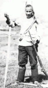 A black-and-white photo of a Native American man standing outdoors, wearing traditional and western clothing, holding a rifle in one hand and resting the other on a long staff.