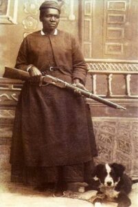 A Black woman wearing a dress and headscarf sits holding a rifle across her lap. A black and white dog lies at her feet. The background features a decorated wall and railing. The image is sepia-toned and historical.