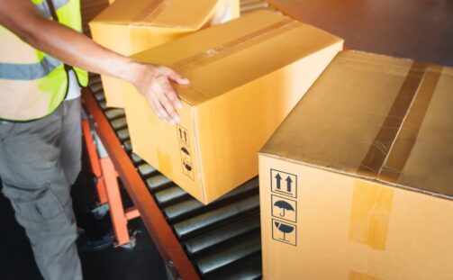 A worker wearing a yellow safety vest moves large cardboard boxes along a conveyor belt in a warehouse. The boxes display handling symbols, indicating they should be kept upright and handled with care.