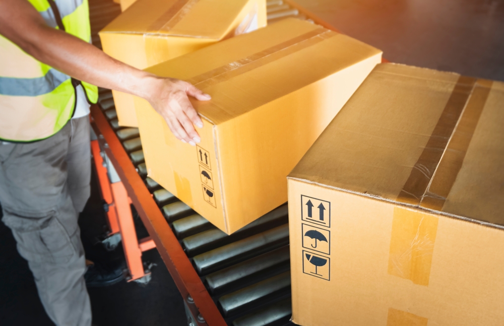 A worker wearing a yellow safety vest moves large cardboard boxes along a conveyor belt in a warehouse. The boxes display handling symbols, indicating they should be kept upright and handled with care.